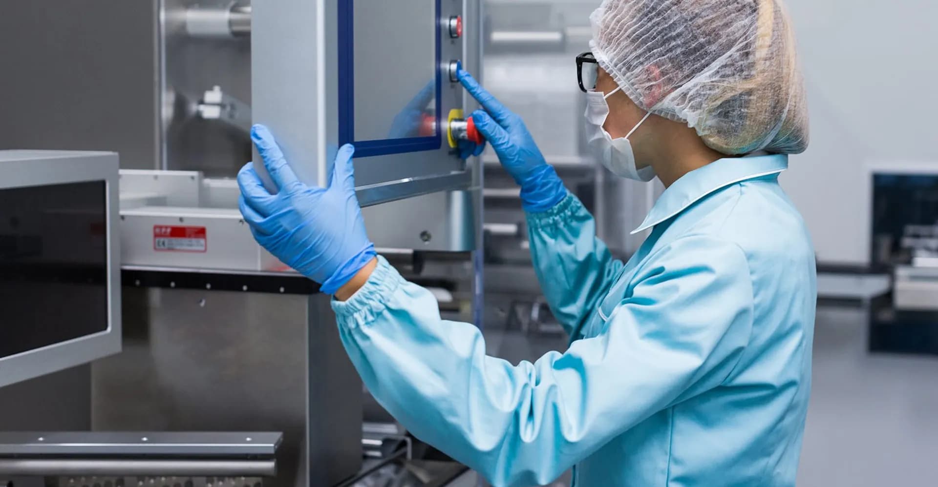 Técnica de touca, máscara e jaleco azul opera o painel de um equipamento de aço inox no laboratório.