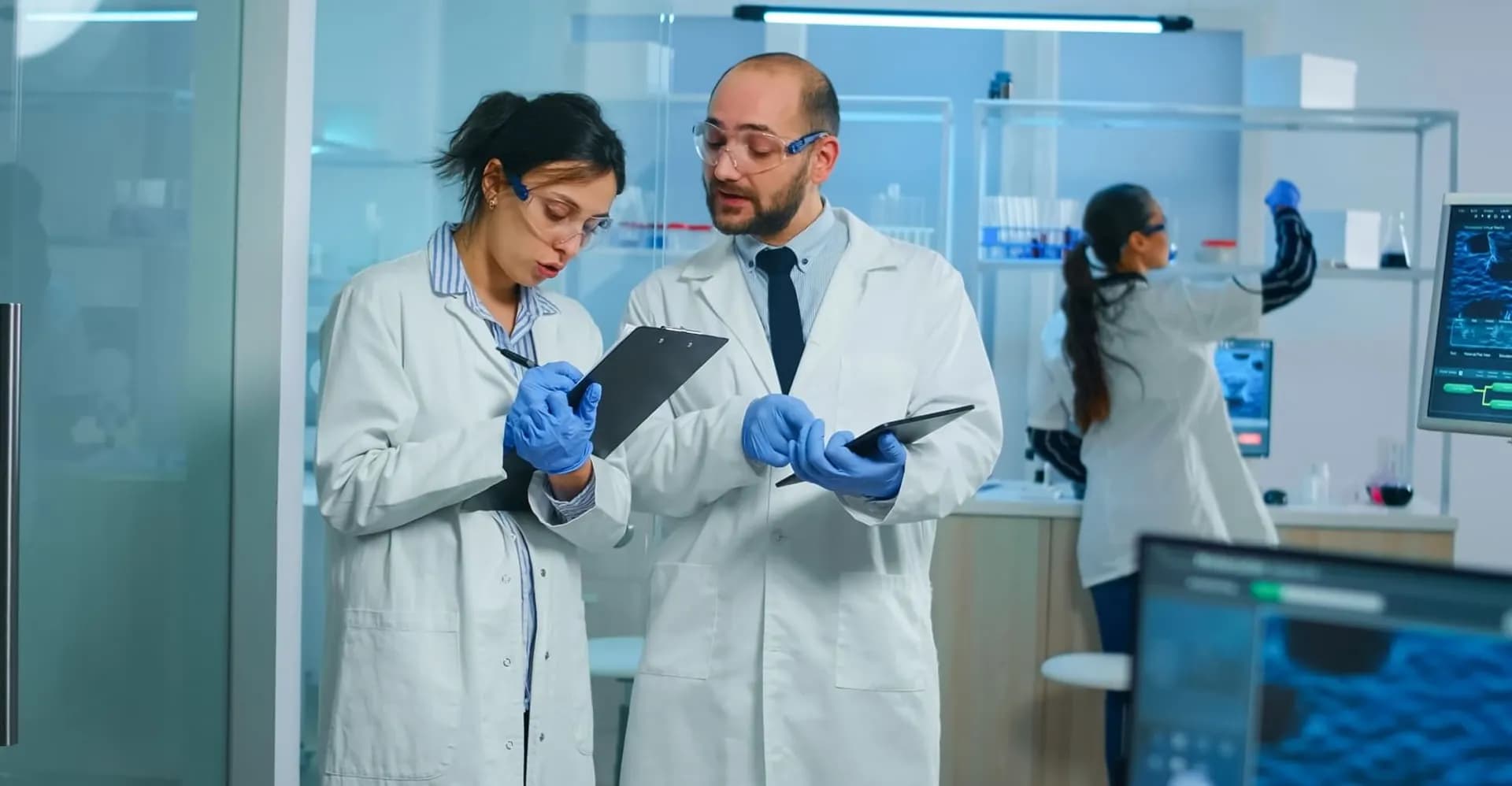 Dois pesquisadores de jaleco revisam anotações em pranchetas num laboratório, enquanto uma terceira pessoa trabalha ao fundo.