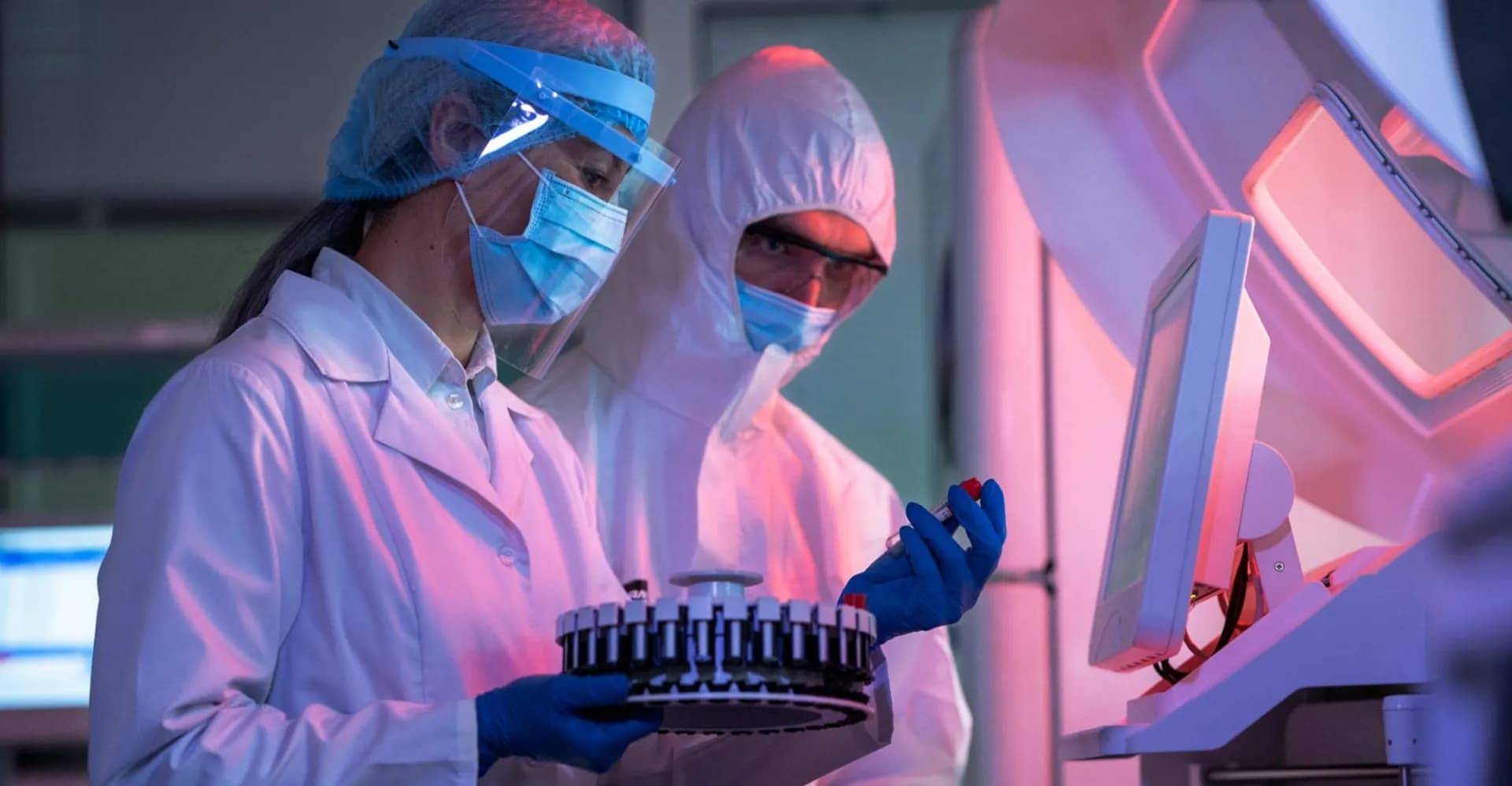 Duas pessoas com máscara, protetor facial e roupa de proteção examinam uma bandeja circular de amostras sob luz rosada.