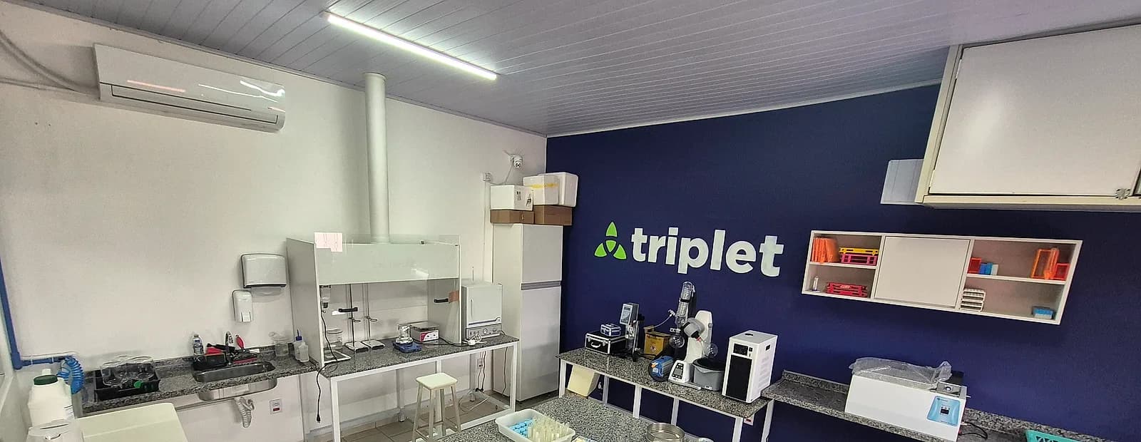 Vista geral do laboratório da Triplet: bancadas de granito com capela de exaustão, agitadores e equipamentos de análise, e a marca aplicada na parede azul.