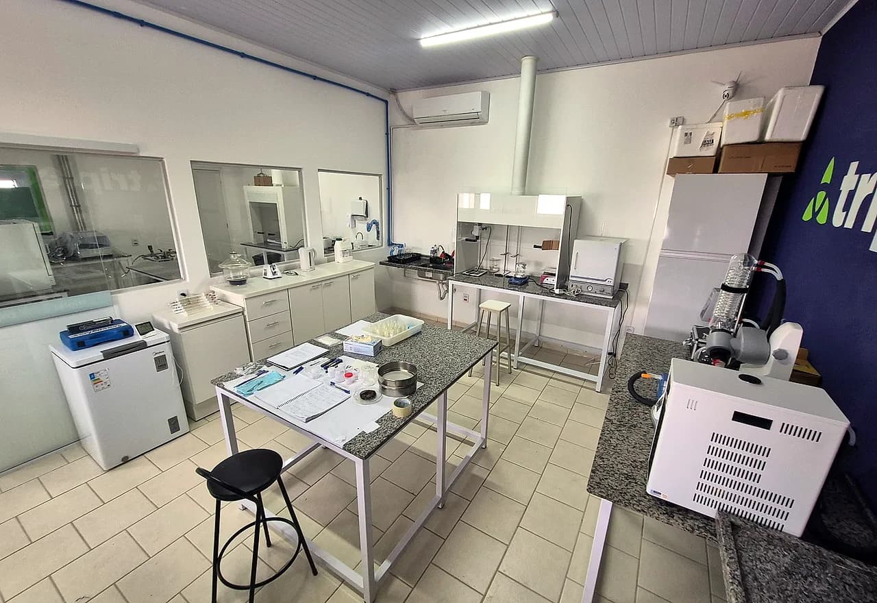 Sala de síntese da Triplet vista da entrada: mesa central de trabalho, freezer, capela de exaustão, evaporador rotativo e a parede azul com a marca.