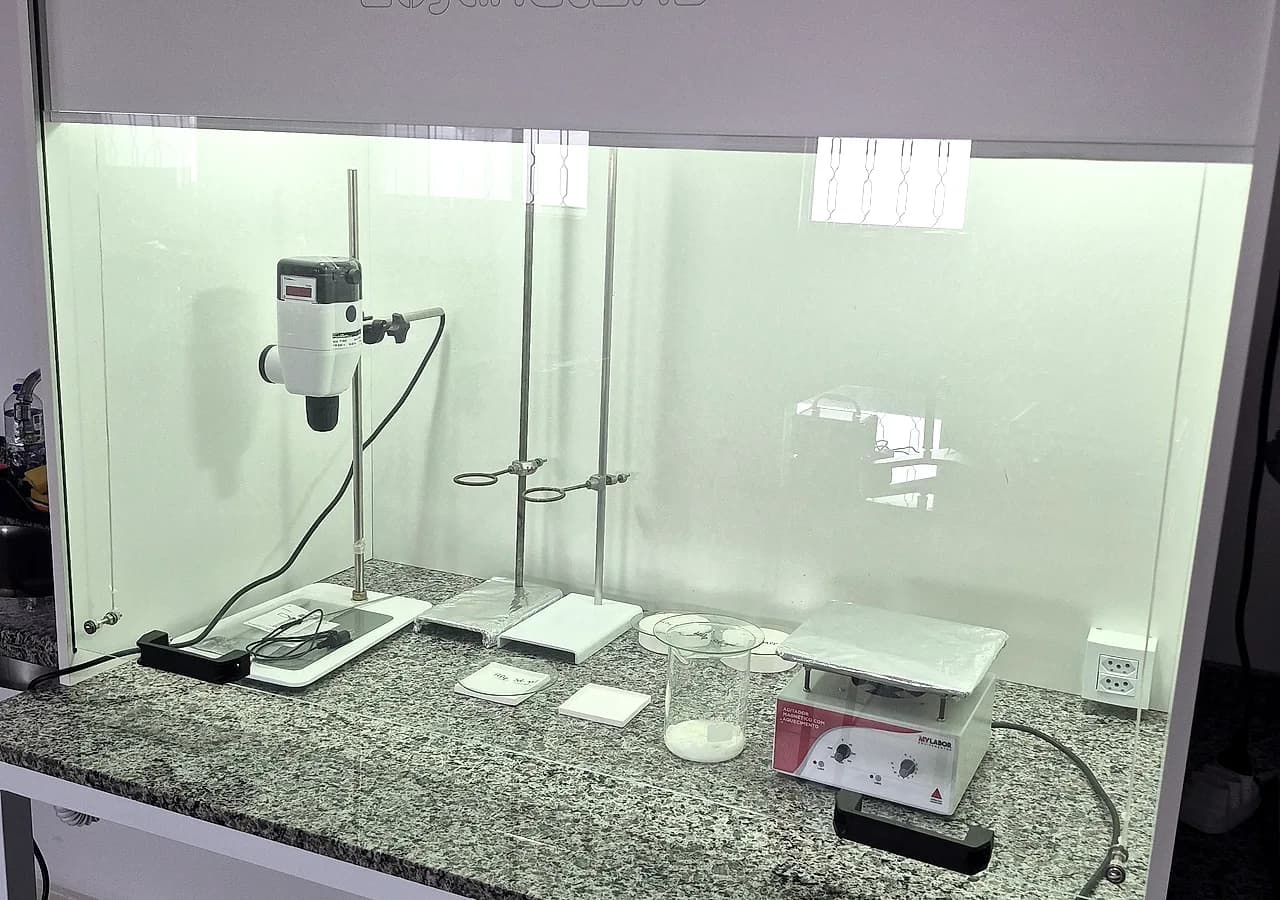 Interior de uma capela de exaustão com agitador mecânico em haste, suportes de vidraria, béquer com pó branco e agitador magnético com aquecimento sobre a bancada de granito.
